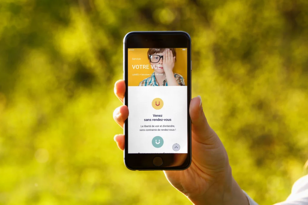 Personne tenant un smartphone affichant une page de services d'opticiens, avec le texte 'Votre vue' et une section invitant les clients à venir sans rendez-vous.