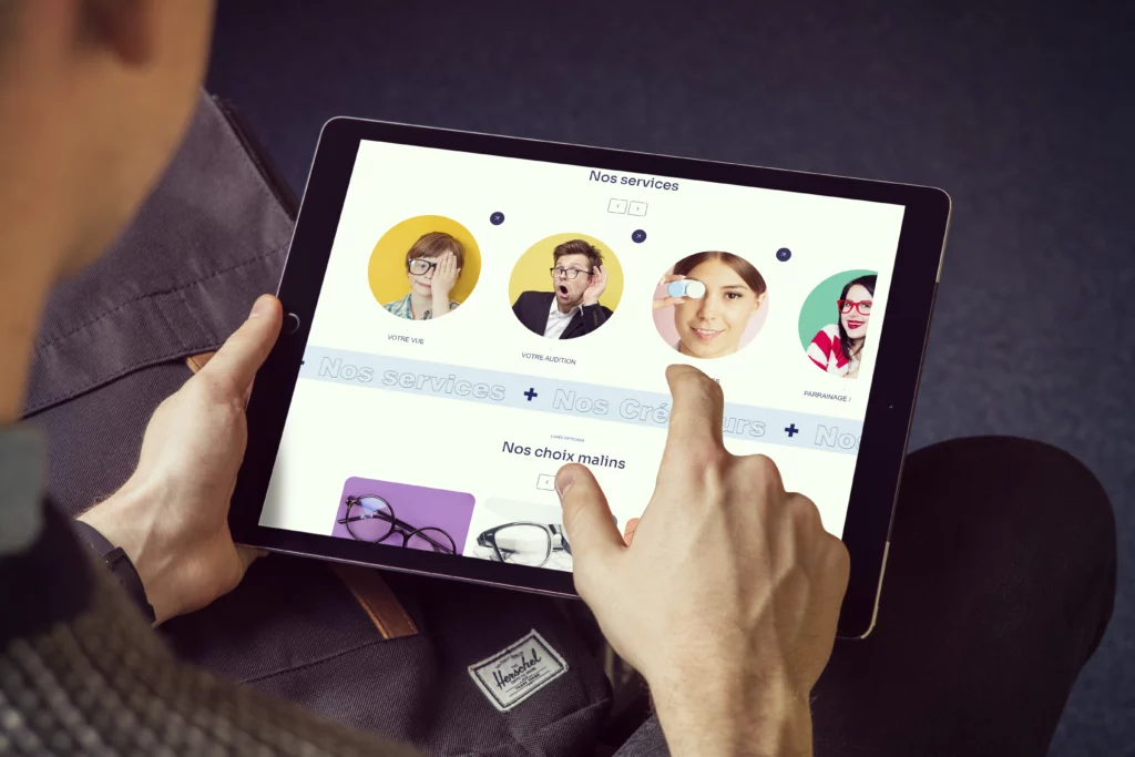 Personne utilisant une tablette pour naviguer sur un site web d'opticiens. L'écran affiche des services tels que votre vue, votre audition, et parrainage, avec des images de personnes portant des lunettes.