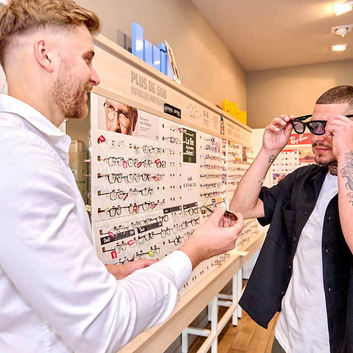 Opticien de Luneo Opticiens aidant un client à essayer des lunettes devant un présentoir de montures